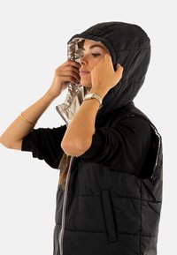 Chaleco acolchado negro con capucha forrada de plata, con cremallera frontal y bolsillos laterales. La tela parece suave y resistente al agua.