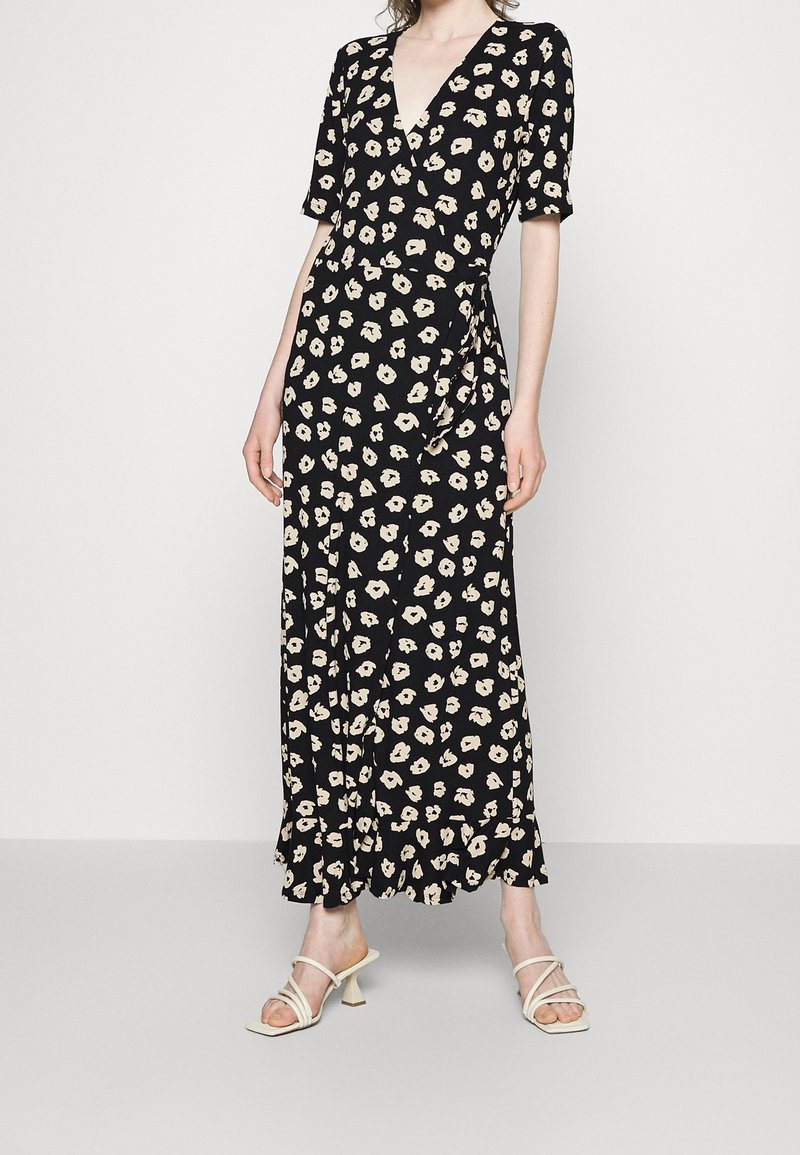 Femme portant une robe portefeuille noire à motif floral blanc et manches courtes, associée à des sandales blanches à talons et à brides.