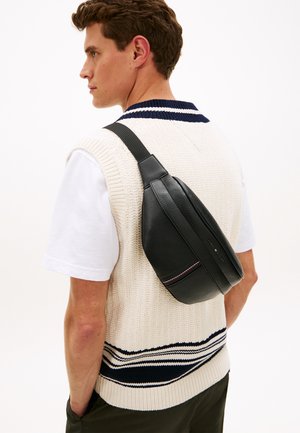 Čierna kožená taška cez rameno s textúrovaným povrchom a kontrastnými červenými a bielymi pruhmi; nosená cez krémovú pletenú vestu.