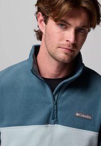 Pullover in fleece di colore verde acqua e azzurro chiaro con colletto a metà zip e patch con logo Columbia sul petto. Texture morbida e design casual.