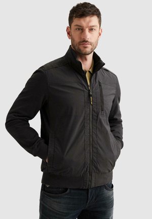 Homme portant une veste noire zippée avec les mains dans les poches, debout devant un fond gris clair uni.