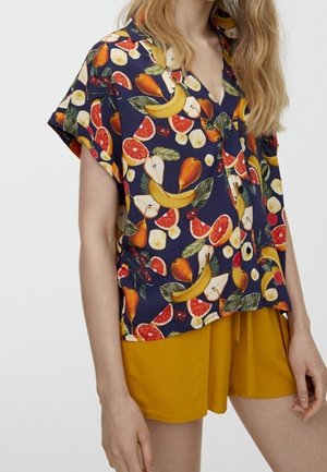 Haut à manches courtes, de couleur bleu marine, avec un motif de fruits vifs comprenant des bananes, des poires et des agrumes. Associé à un short jaune uni. Tissu lisse.