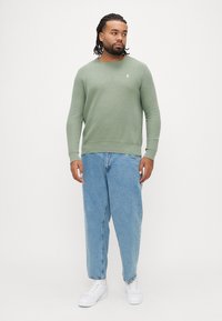 Πράσινο πλεκτό πουλόβερ με στρογγυλή λαιμόκοψη, συνδυασμένο με χαλαρά μπλε τζιν και λευκά αθλητικά παπούτσια, με μικρό λογότυπο στο στήθος.