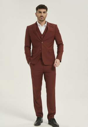 Homme debout les mains dans les poches, portant un costume ajusté rouge foncé, une chemise blanche et des chaussures habillées noires, sur un fond uni.