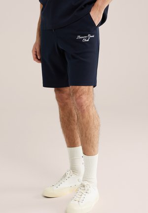 Muškarac u tamnoplavim kratkim hlačama s natpisom „Leisure Court Club“, bijelim čarapama i bijelim tenisicama na vezanje, stoji s jednom rukom u džepu.