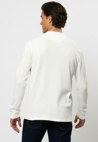 Mand med mørkt hår iført en hvid langærmet waffle-strikket trøje og mørke jeans, set bagfra mod en ensartet baggrund.