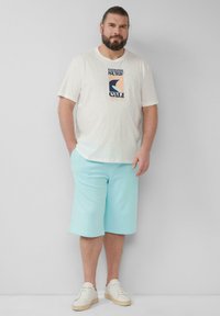 T-shirt crème avec un design graphique noir et pastel, associée à un short bleu clair. Des baskets blanches complètent la tenue. Style décontracté.
