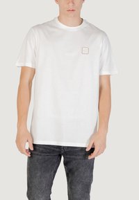 T-shirt bianco in cotone con maniche corte e collo tondo. Presenta una piccola toppa con logo quadrato sul petto. Indossato con jeans grigio scuro.