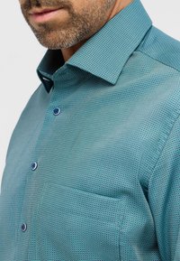 Turkis mønstret skjorte med en struktureret krave. Har små runde knapper og en brystlomme, lavet af et glat stof.