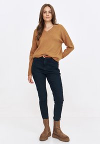Brązowy sweter z dekoltem w serek, granatowe dżinsy i beżowe botki za kostkę. Sweter ma prążkowany dekolt; dżinsy są dopasowane. Botki mają teksturowane podeszwy.
