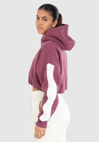 Zkrácená mikina v barvě mauve s kapucí, bílé pruhy na rukávech a měkká textura. Vytvořeno v kombinaci se svetle barevnými kalhotami.