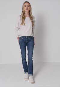 Světlé šedé pletené svetr, slim-fit tmavě modré džíny a bílé tenisky. Model stojí na jednobarevném bílém pozadí.