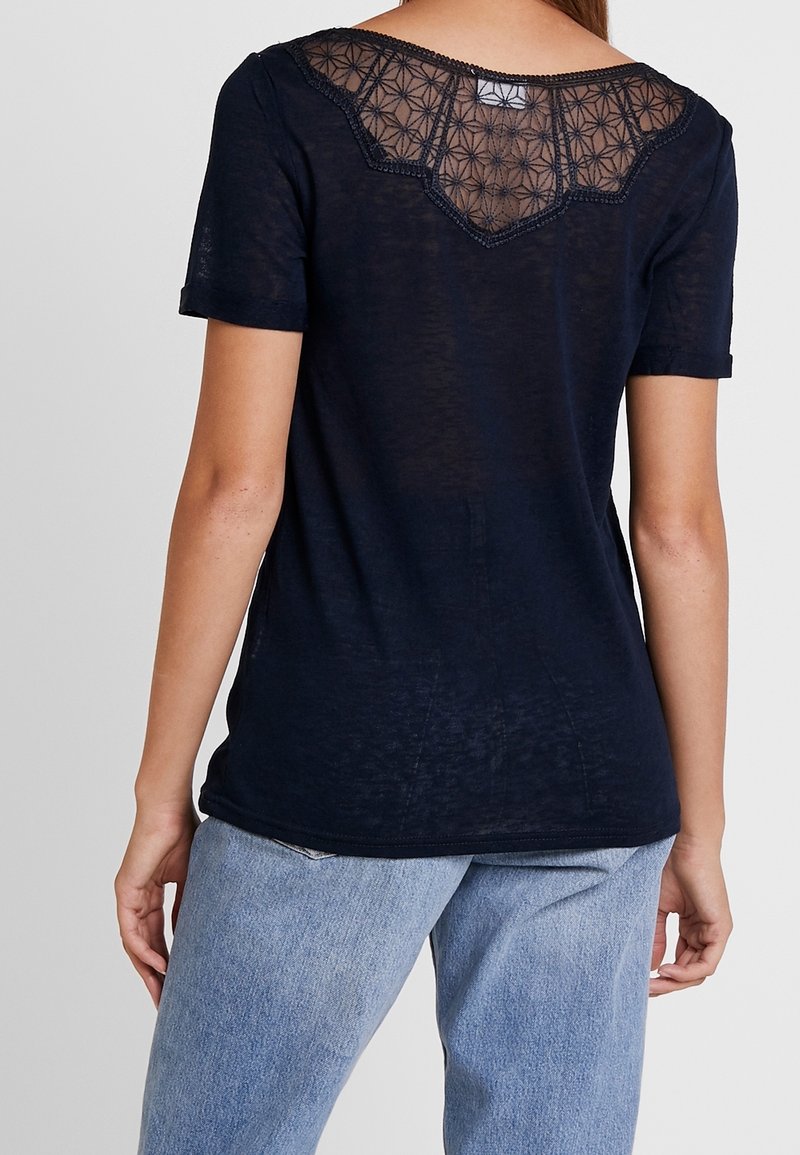 Femme portant un haut noir à manches courtes avec une empiècement en dentelle florale transparente sur le haut du dos, associée à un jean bleu clair, debout de dos.