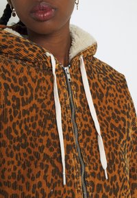 Huppari, jossa on ruskea ja musta leopardi-tulostus, valkoinen sherpa-vuori, vetoketjukiinnitys ja nyöri hopeisilla yksityiskohdilla. Teksturoitu kangas.