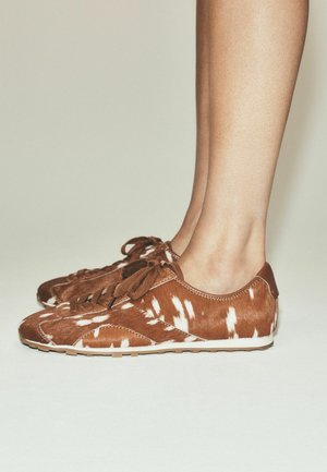 Brune og hvide sneakers med pletter og snørebånd båret på bare fødder mod en ensfarvet lys baggrund.