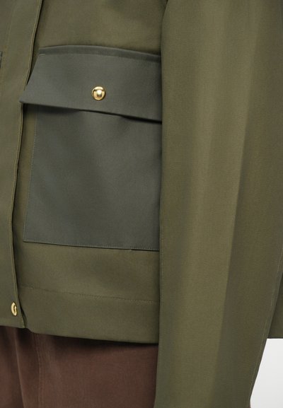 Olive green jacket with a smooth texture, featuring a large front pocket secured by a gold snap button and a simple straight cut.