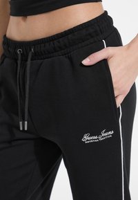 Black sweatpants with an elastic waistband and drawstring. Features white side stripes and a "Guess Jeans" logo in white embroidery.