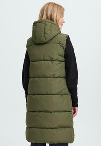 Olivgrüne Steppweste mit Kapuze, die horizontale Steppnähte und ein langes Design aufweist. Darunter ist ein schwarzes Langarmshirt sichtbar.