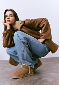 Botines de gamuza marrón con una suela texturizada y costuras en zigzag, combinados con jeans azules de corte ancho y un abrigo marrón forrado con borrego.