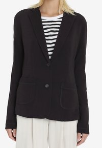 Blazer nero con colletto a revers, due tasche frontali e chiusura a bottone. Tessuto morbido con dettagli di cucitura ben visibili. Indossato sopra una camicia a righe.