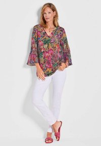Bluse mit Blumenmuster und Glockenärmeln in lebhaften Farben, kombiniert mit weißen Hosen und rosafarbenen Sandalen. Leichter, luftiger Stoff mit V-Ausschnitt.