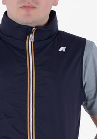 Mornarska brezrokavnika s visokim ovratnikom, sprednjim zadrgo z večbarvnimi črtami in majhnim logotipom na levi prsih. Gladka tekstura.