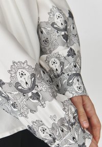Blusa blanca con un patrón de encaje floral en blanco y negro en las mangas, con una textura suave y detalle de puño fruncido.