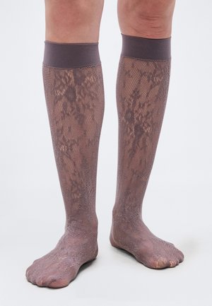 Beine mit kniehohen, durchsichtigen braunen Spitzensocken mit festem Bündchen, die nackte Zehen und ein leichtes Blumenmuster im Netzstoff zeigen.