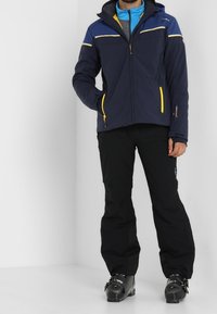 Chaqueta de esquí azul marino con acentos amarillos y azules, bolsillos con cremallera y capucha; combinada con pantalones de esquí negros y botas. Textura suave.