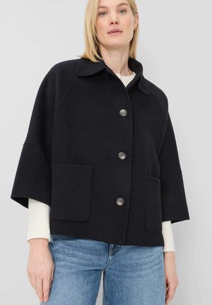 Frau trägt einen schwarzen Knopf-Mantel mit weiten Ärmeln und aufgesetzten Taschen über einem weißen Pullover und blauen Jeans vor neutralem Hintergrund.