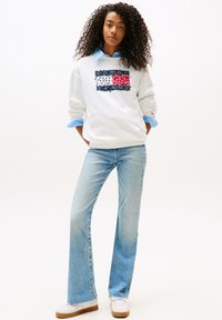 Sudadera blanca con logo gráfico en azul, rojo y navy, combinada con jeans acampanados de color azul claro y zapatillas blancas con suelas de goma.