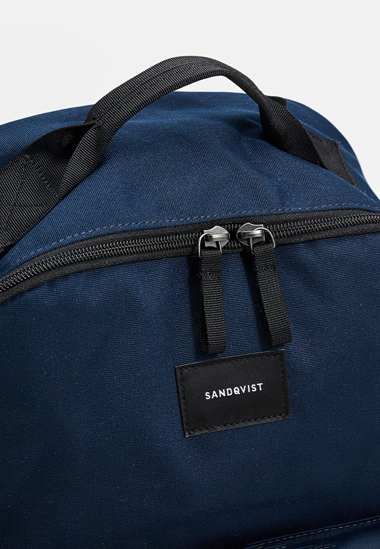 Mochila azul marino con detalles negros, que cuenta con una cremallera y un asa resistentes, material de tela y una etiqueta de marca rectangular negra.