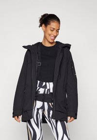 Black hooded parka with front zip; features zipper pockets and a textured finish. Paired with striped pants in black, white, and beige.