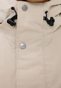 Prenda exterior beige con cierre frontal de cremallera, botones de presión metálicos y cordones ajustables. El material parece liso y ligeramente brillante.