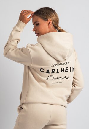 Bež pulover s kapuco z velikim potiskanim logotipom na hrbtni strani, na katerem piše "Copenhagen CARLHEIN Denmark Established 2017." Mehka, teksturirana tkanina.