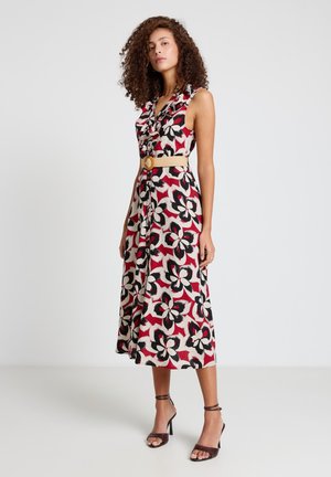 Donna con capelli ricci che indossa un vestito rosso senza maniche con grande stampa floreale nera e bianca, cintura beige e sandali con tacco marroni.