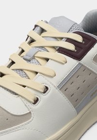 Gros plan d'une basket avec des lacets beige, des panneaux blancs et gris, un liseré œillet bordeaux et des détails latéraux perforés.