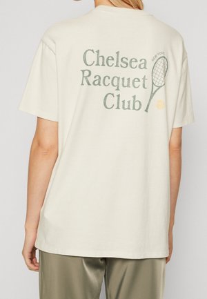 Kobieta w białej koszulce z napisem "Chelsea Racquet Club", ilustracją rakiety tenisowej i piłki na plecach.