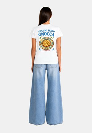 Donna con capelli lunghi e scuri che indossa una t-shirt bianca con l'illustrazione di un piatto di gnocchi e testo, abbinata a jeans a gamba larga di colore azzurro chiaro.