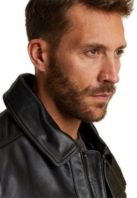 Moški s kratkimi rjavimi lasmi in brado, oblečen v črno usnjeno jakno z zavihanimi ovratniki, obrnjeno desno proti enotni podlagi.