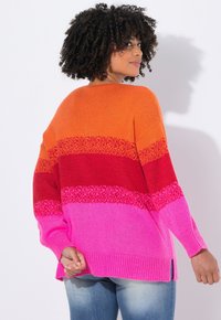 Donna con capelli ricci che indossa un maglione a strisce arancione, rosso e rosa, in piedi e volto di spalle contro uno sfondo bianco.