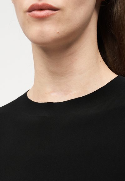 Κοντινή λήψη του κάτω μέρους του προσώπου και του λαιμού γυναίκας που φοράει μαύρη μπλούζα με στρογγυλή λαιμόκοψη, με απλό ανοιχτόχρωμο φόντο.