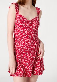 Punainen kukallinen mekko, jossa on tiukka yläosa ja leveä hame, jossa on pieniä valkoisia kukkia, leveät olkaimet ja pyöreä kaula-aukko.