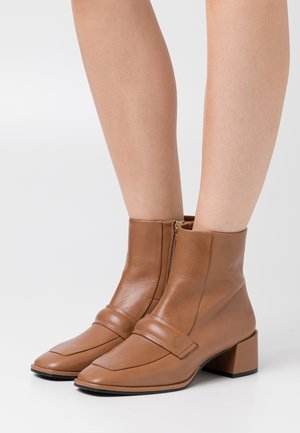 Braune Leder-Stiefeletten mit Blockabsatz, quadratischer Zehenpartie, seitlichem Reißverschluss und einem dekorativen Riemen über der Vorderseite. Glatte Textur und minimalistisches Design.