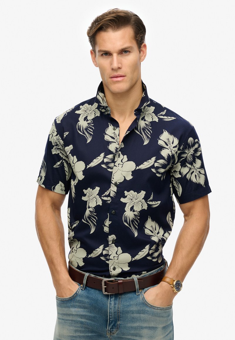 Camisa de manga corta azul marino con un ligero patrón floral, cuello abotonado y tejido de algodón suave; combinada con jeans azul claro y cinturón marrón.