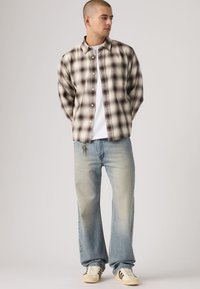 Πουκάμισο με κουμπιά σε μπεζ και καφέ καρό, συνδυασμένο με ανοιχτόχρωμο μπλε τζιν με χαλαρή εφαρμογή. Το μοντέλο φοράει λευκό μπλουζάκι και μπεζ αθλητικά παπούτσια.