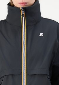 Črna jakna z visokim ovratnikom, ki ima večbarven zadrgo in logo. Material je gladek in lahek ter ima sproščen kroj.