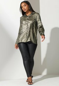 Camicia lunga a maniche con chiusura a bottoni di un verde oliva metallico con una texture morbida. Abbinata a leggings neri eleganti e tacchi a strappi neri.