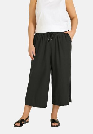 Kvinde iført sorte, vide bukser med snøre i taljen, hvid ærmeløs top og sorte sandaler med åbne tæer, stående med den ene hånd i lommen.