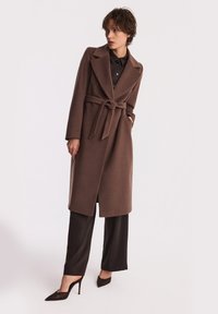Brązowy płaszcz z wełny z paskiem w talii, z wykładanym kołnierzem i długimi rękawami, w połączeniu z czarnymi szerokimi spodniami i spiczastymi obcasami.